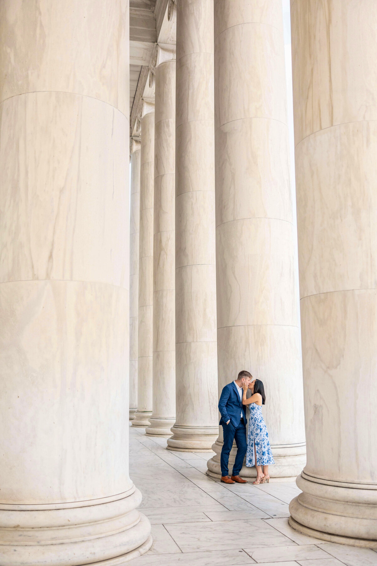 A couple stands closely between towering marble columns, sharing an intimate moment. The man is in a blue suit and the woman in a floral dress. The setting suggests a neoclassical architecture style.