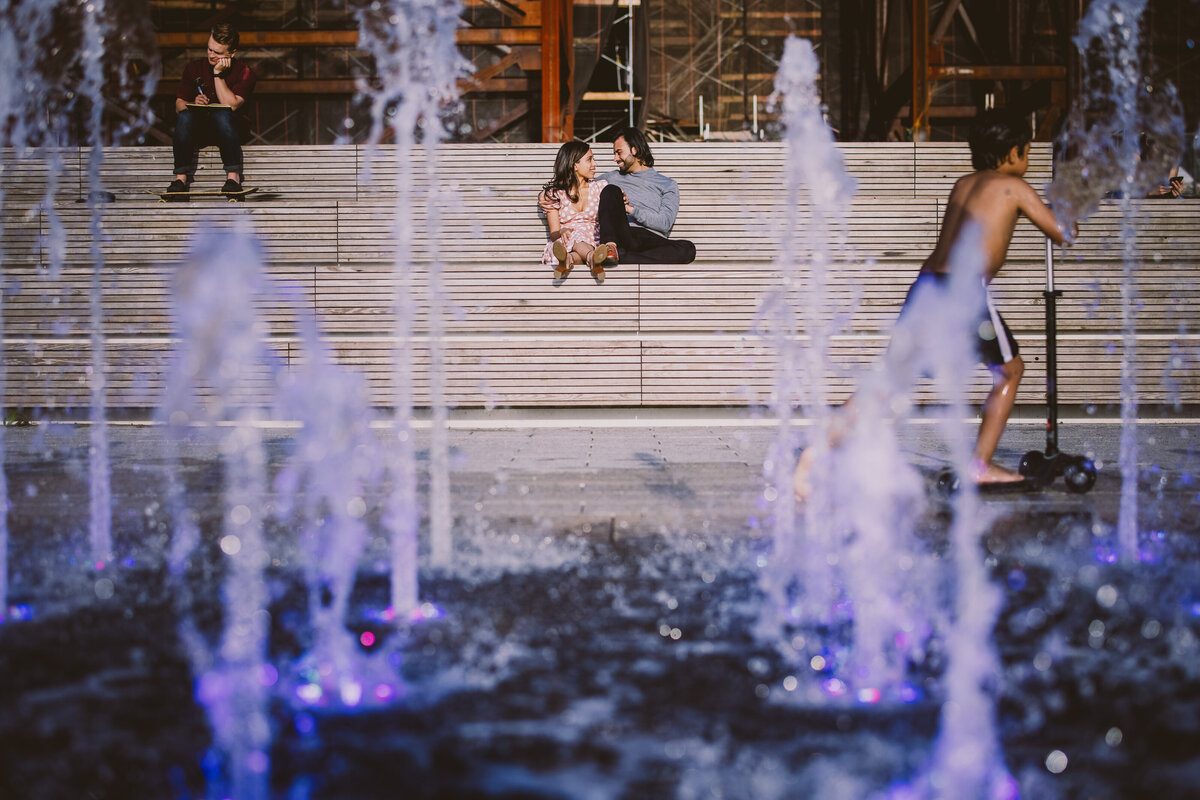An engaged couple sitting on the bleachers at domino park with the water fountains in the foreground