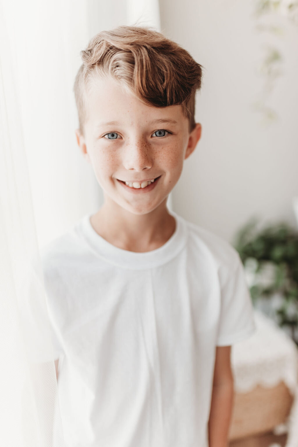 boy standing near window wearing white shirt with plants in the background