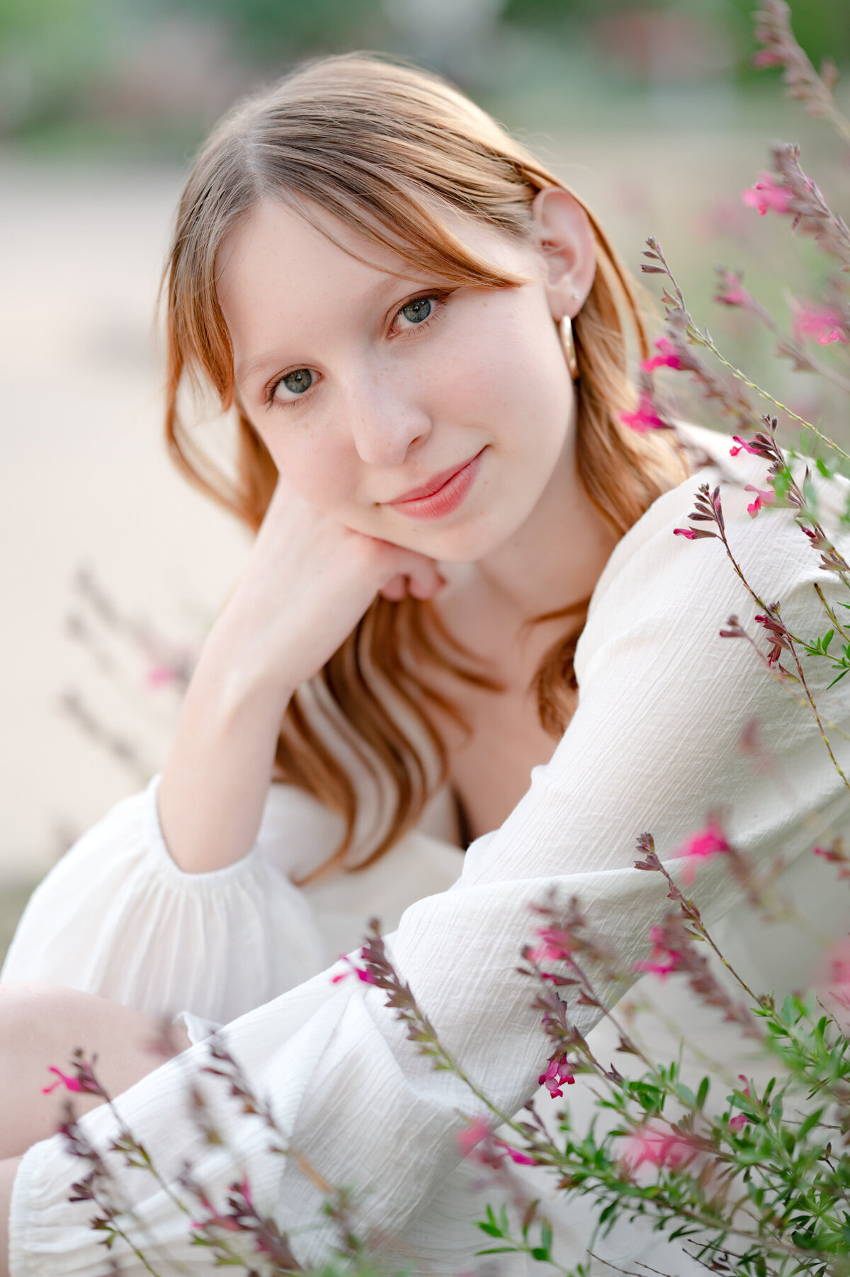 Senior picture of a red-headed girl in a white dress sitting among pink flowers.