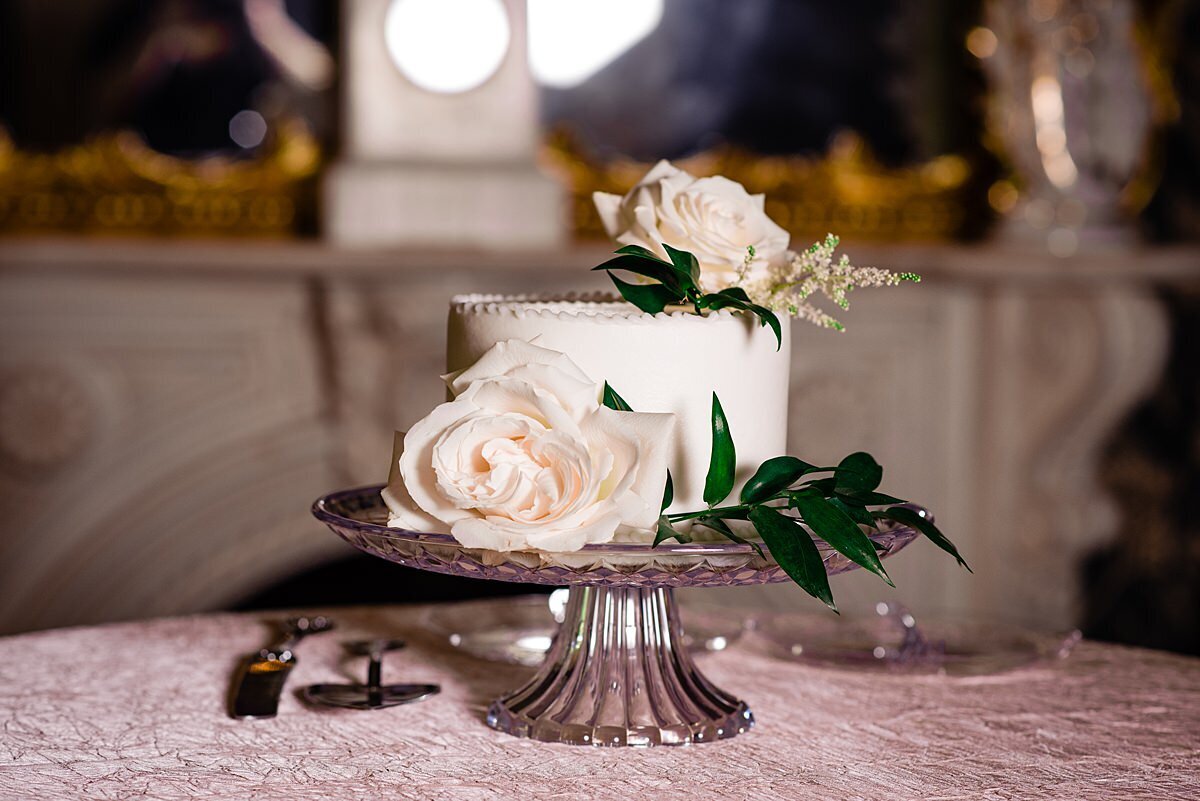 A small one tier wedding cake on a silver footed platter with a cake knife and server , plate and fork on the table below. The cake stand is sitting on a blush table cloth with a white mantle in the background.