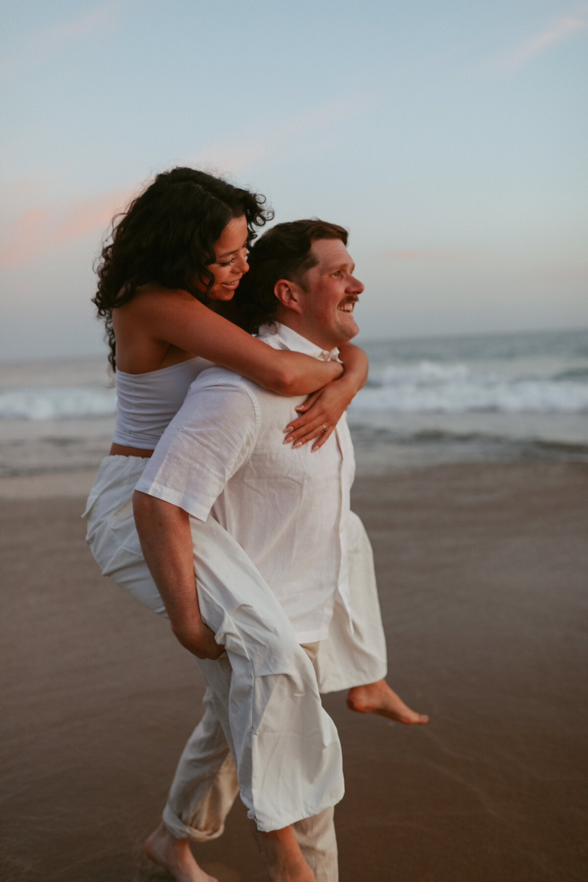 engagement photos at the beach , guy carrying his girlfriend piggy back style down the beach