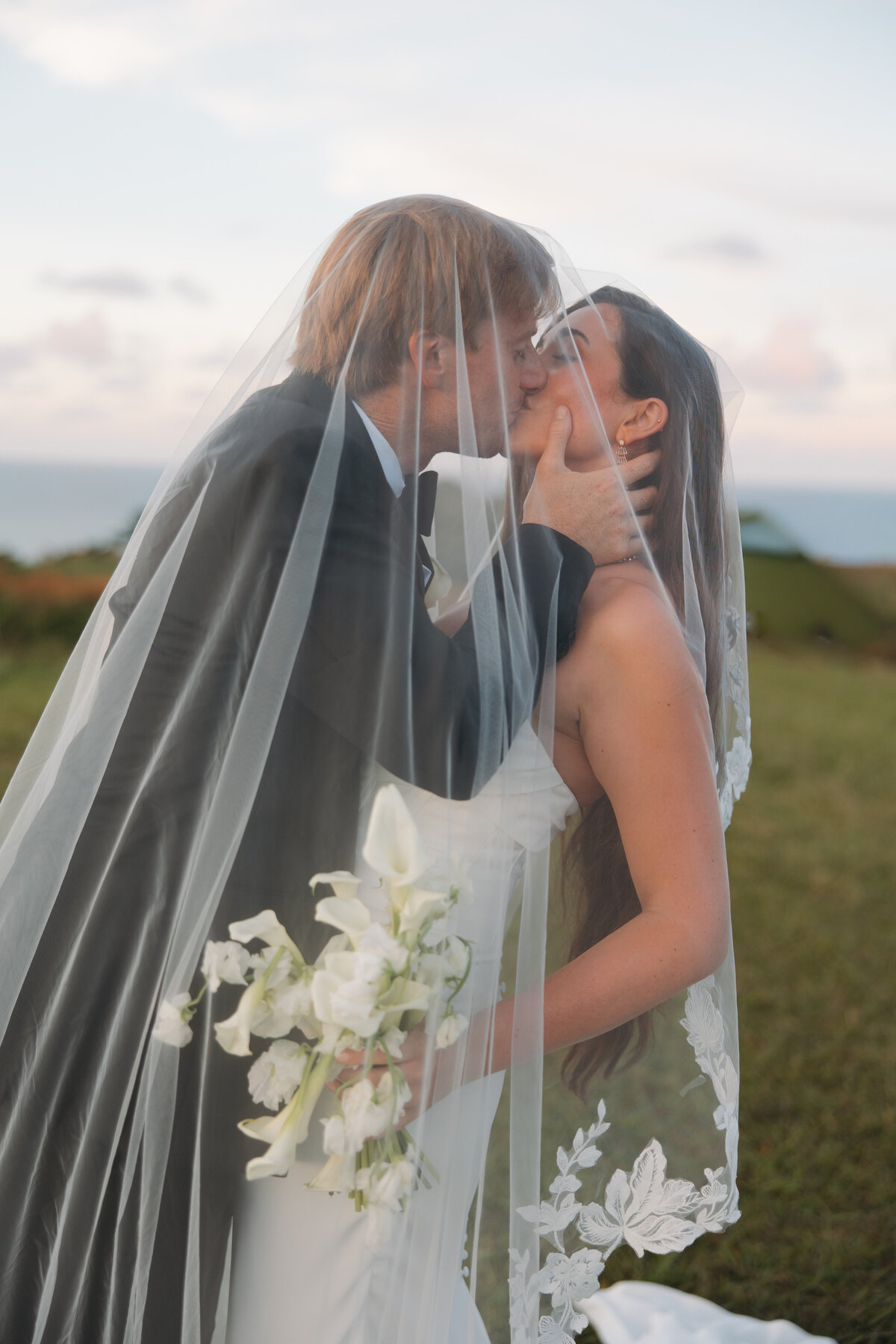 Bride and groom share a romantic kiss under the veil at North Shore Preserve in Kauai, with soft ocean views and pastel skies in the background.