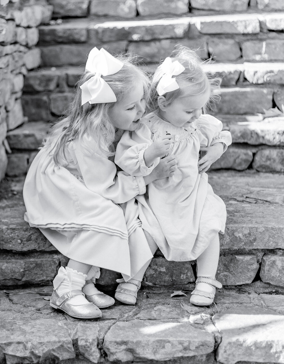 Black and white portrait of two young sisters sitting together on stone steps, dressed in classic smocked dresses with matching white bows. The older sister leans in with a smile, capturing the timeless beauty of sibling bonds in a heartfelt Birmingham family photography session.