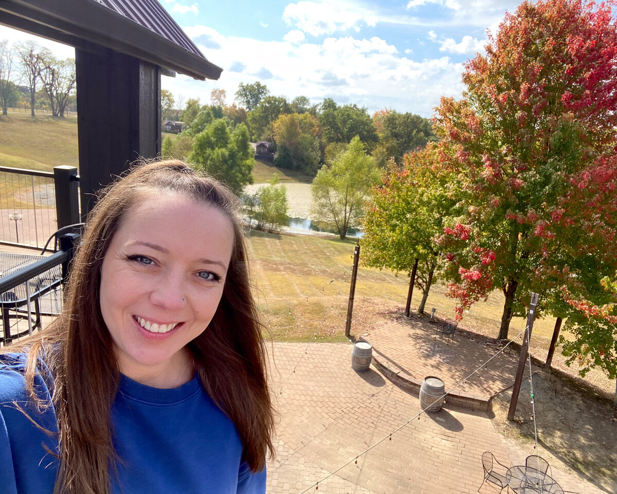 woman with long dark hair standing on a deck, with a pond and trees in the background, smiling at the camera