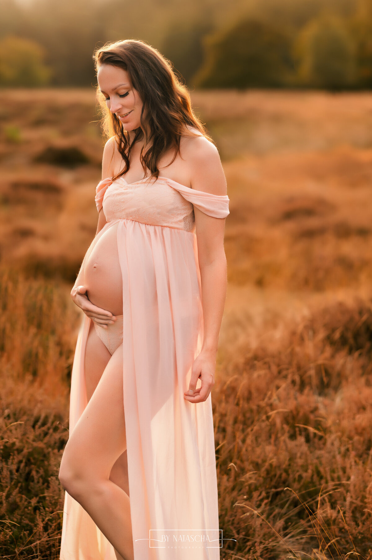 Zwangere vrouw met haar ene arm langs het lichaam en de andere hand houdt haar baby buik vast vanaf de onderzijde en ze kijkt naar haar zwangere buik in de heide van Herperduin