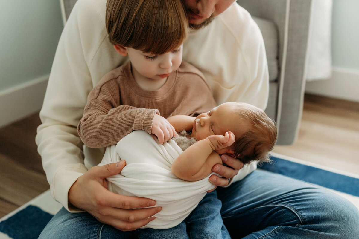A toddler boy sits on his dad's lap while holding his newborn baby brother