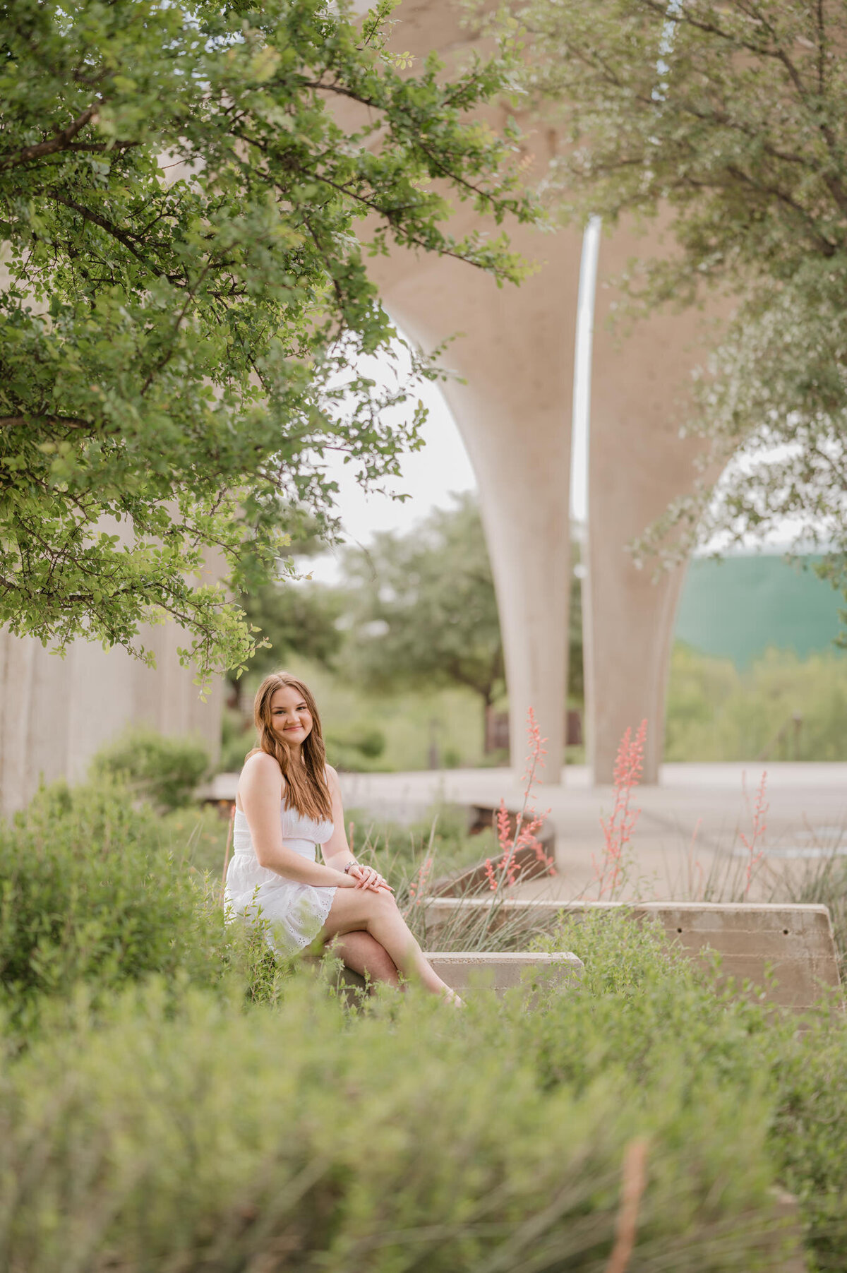 Senior picture of a girl in a white dress sitting surrounded by greenery with concrete arches in the background.