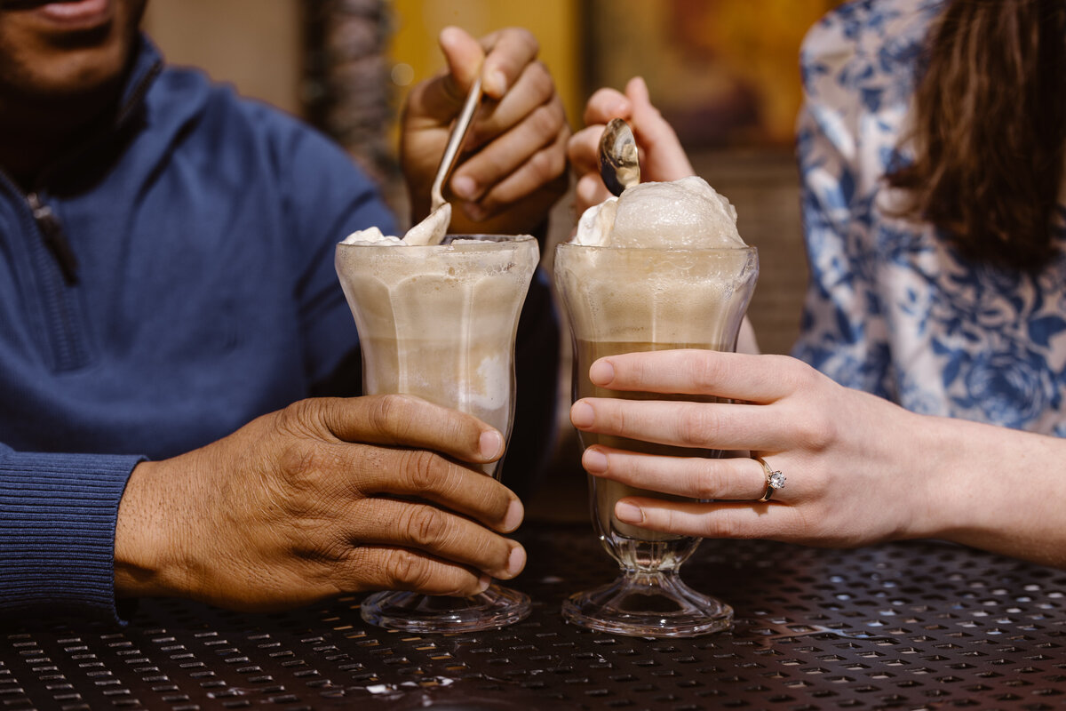 Close-up of an engaged couple enjoying root beer floats at an outdoor table, highlighting the woman’s engagement ring and the shared moment.