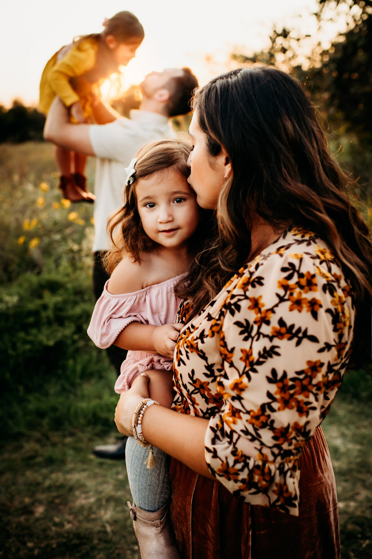 Family Photographer, A mother kisses her young daughter as dad lifts other daughter into the air