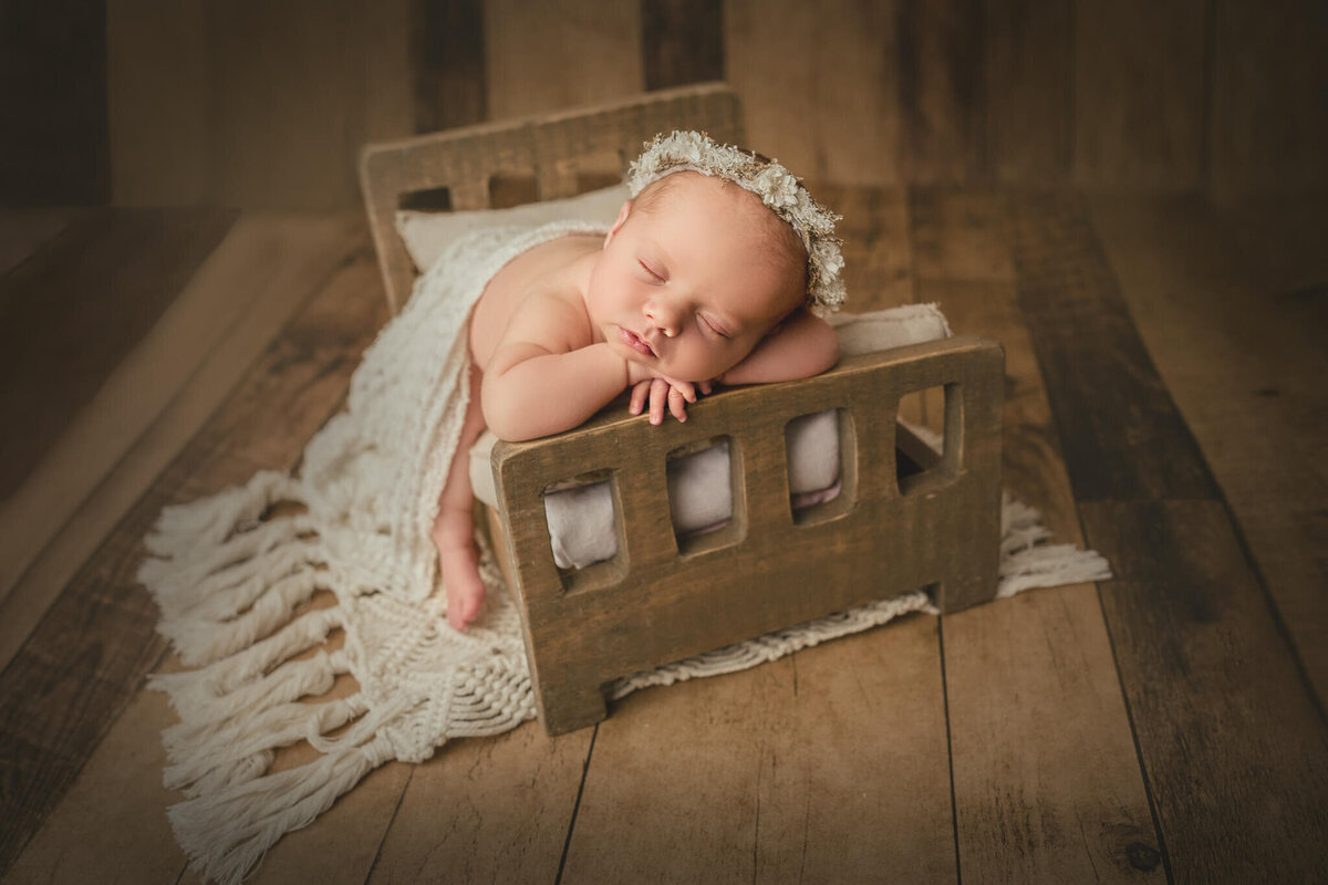 newborn baby girl poses with chin on hands in tiny wooden bed