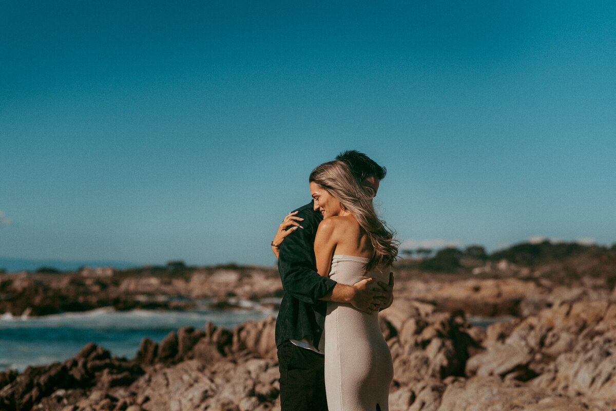 Newly engaged couple hugging on the rocky shoreline at Asilomar State Beach in Pacific Grove