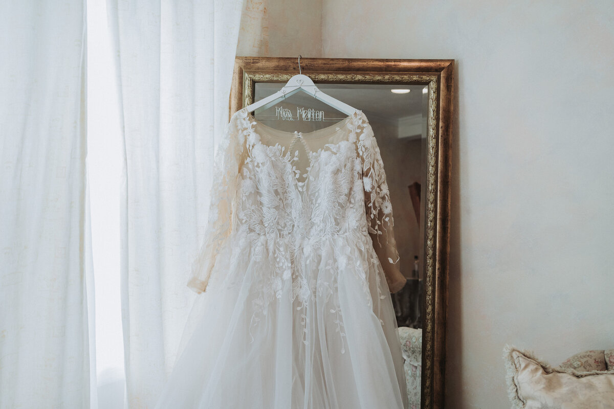 An elegant long-sleeved lace wedding dress hanging on a custom "Mrs. (Name)" hanger, reflected in a large gold-framed mirror, showcasing bridal details before the ceremony, photographed by Quinn Photography LLC.