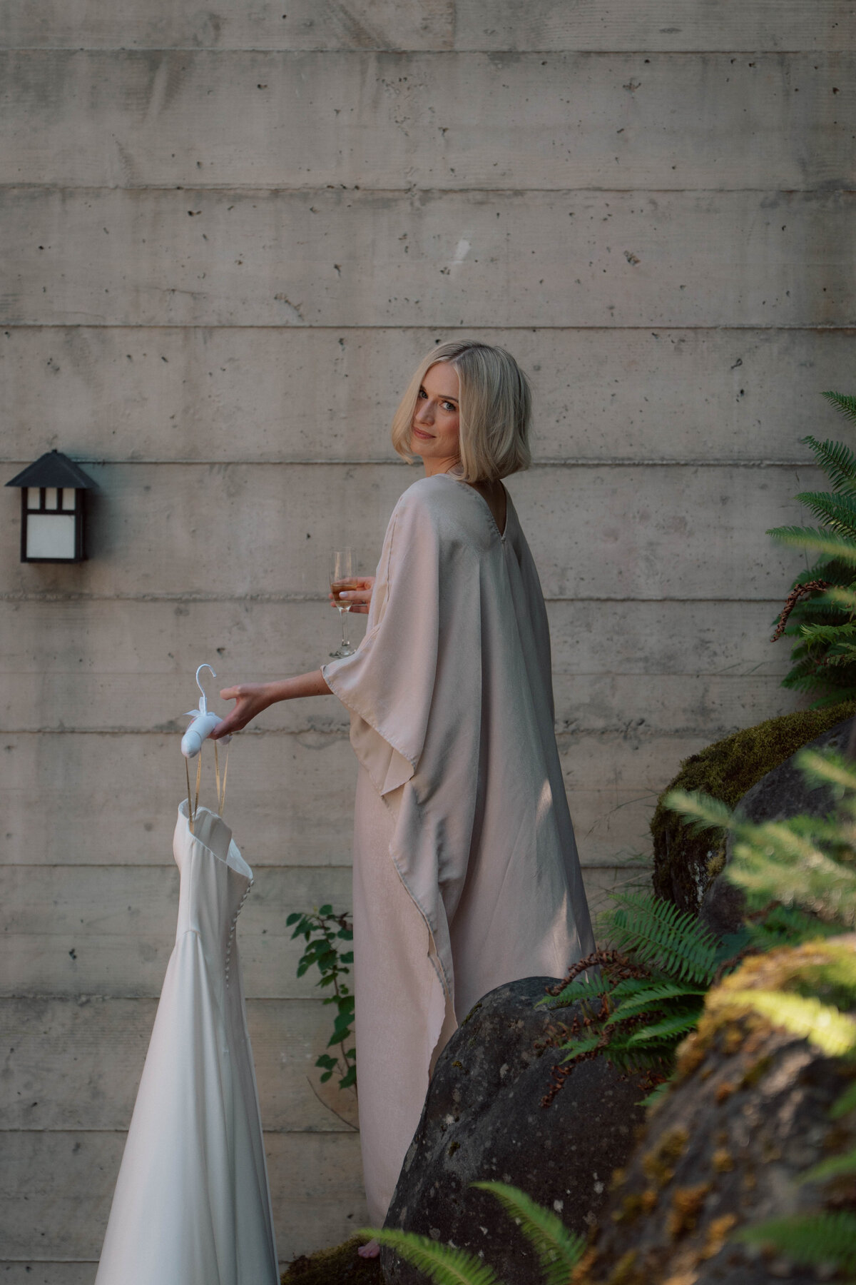 Bride in soft robe holding champagne and wedding gown outdoors against stone wall