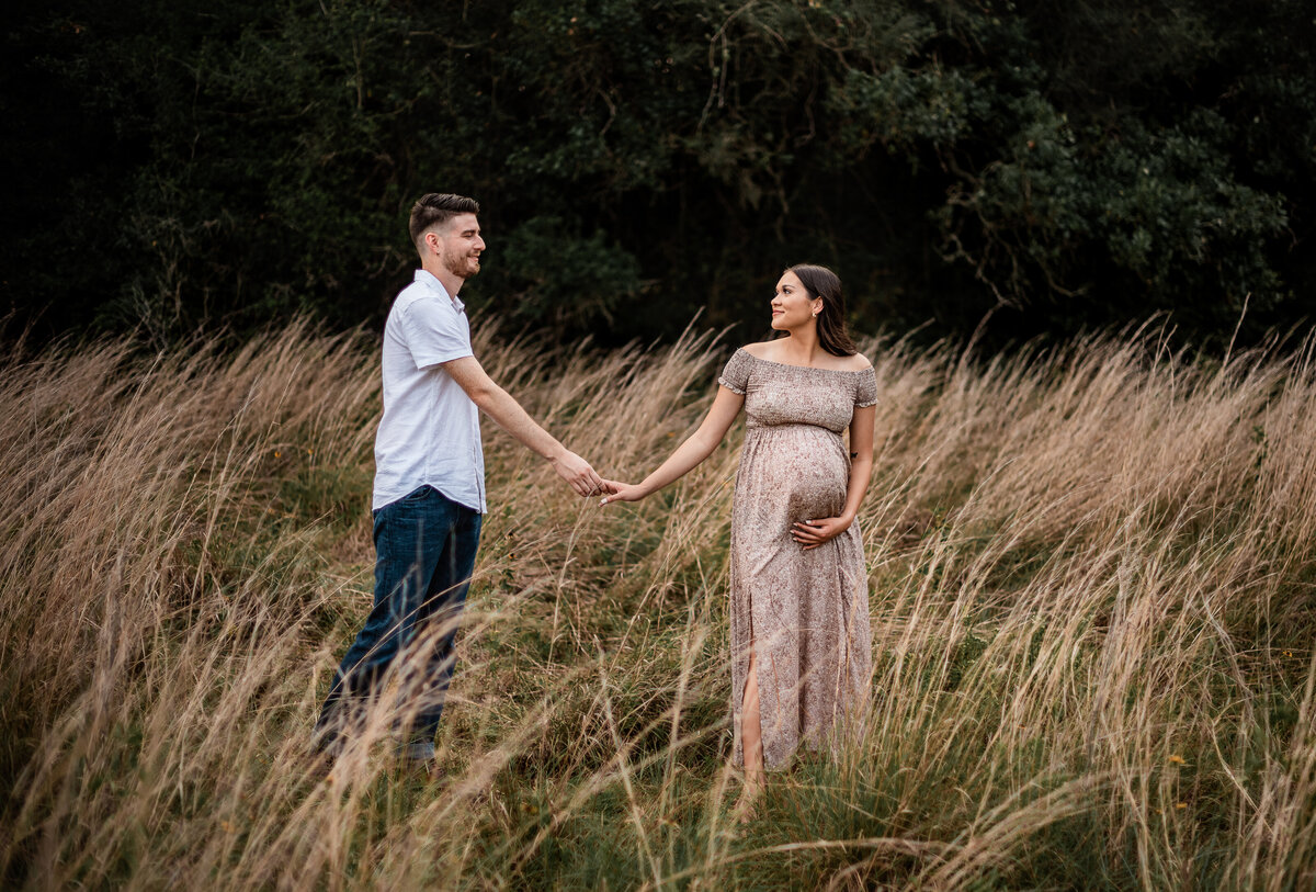 At Brazos Bend State Park, a couple holds hands and walks through a field while the woman holds her baby bump.