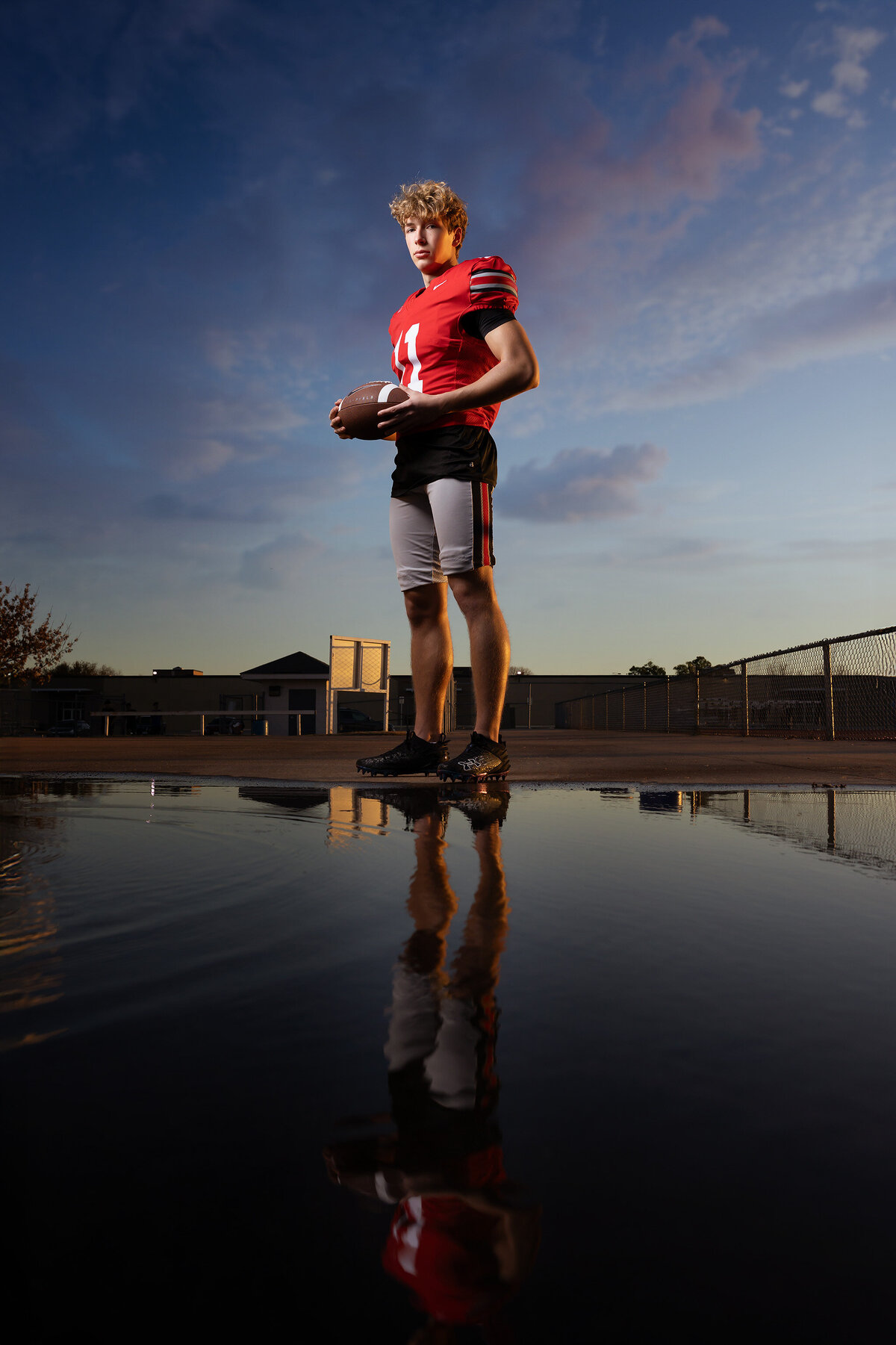 A senior football player stands proudly, with his reflection captured by a Dallas senior photographer during sunset.