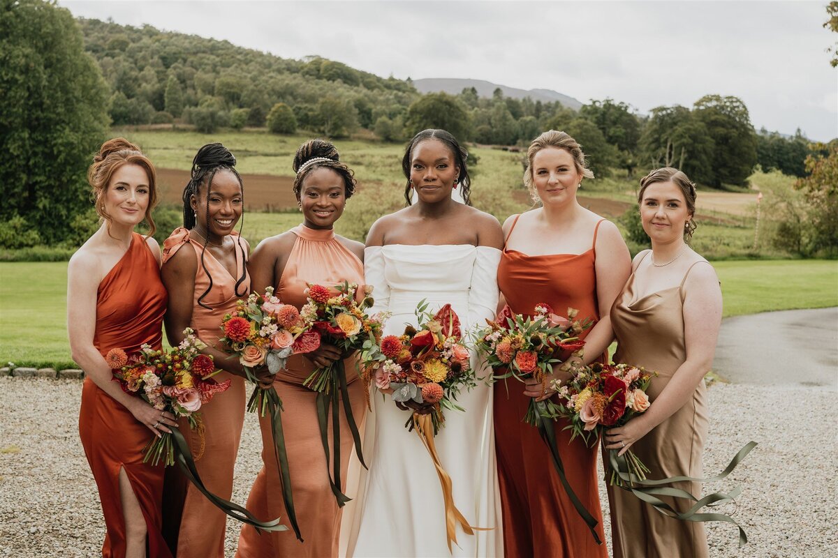A wide photograph of a bride and her bridesmaids. They all hold bouquets and wear autumnal colours. The Aberdeen countryside is seen in the background.