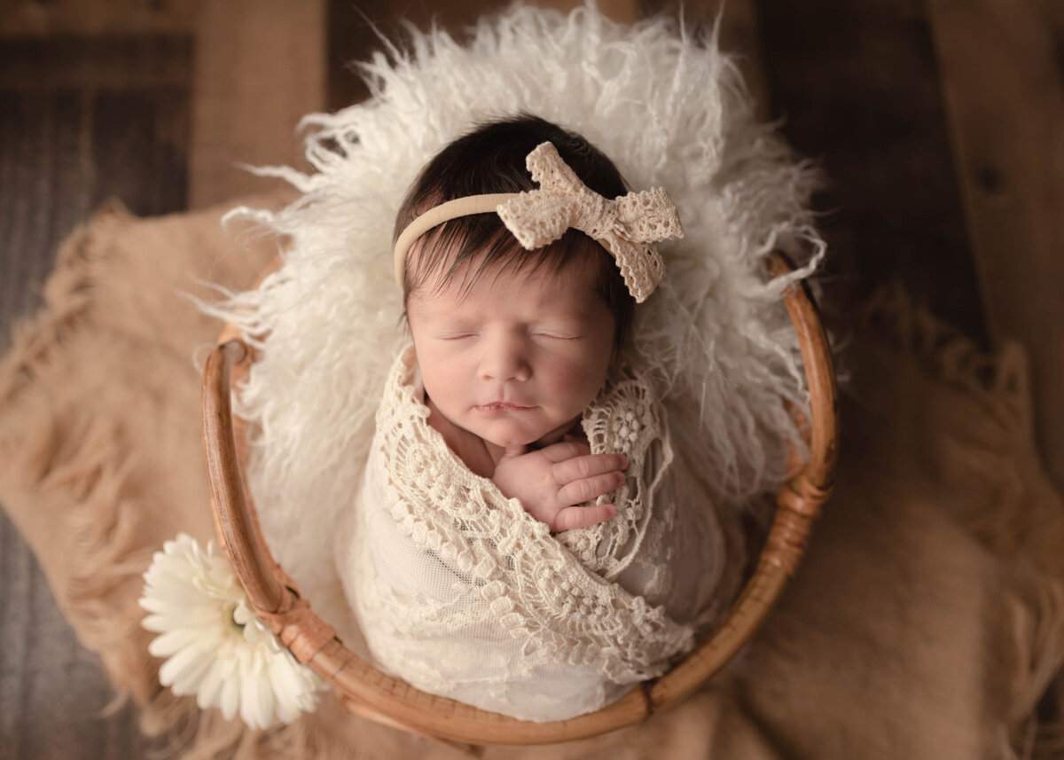 Baby sleeping peacefully in lace wrap with cream headband in basket on a flokati