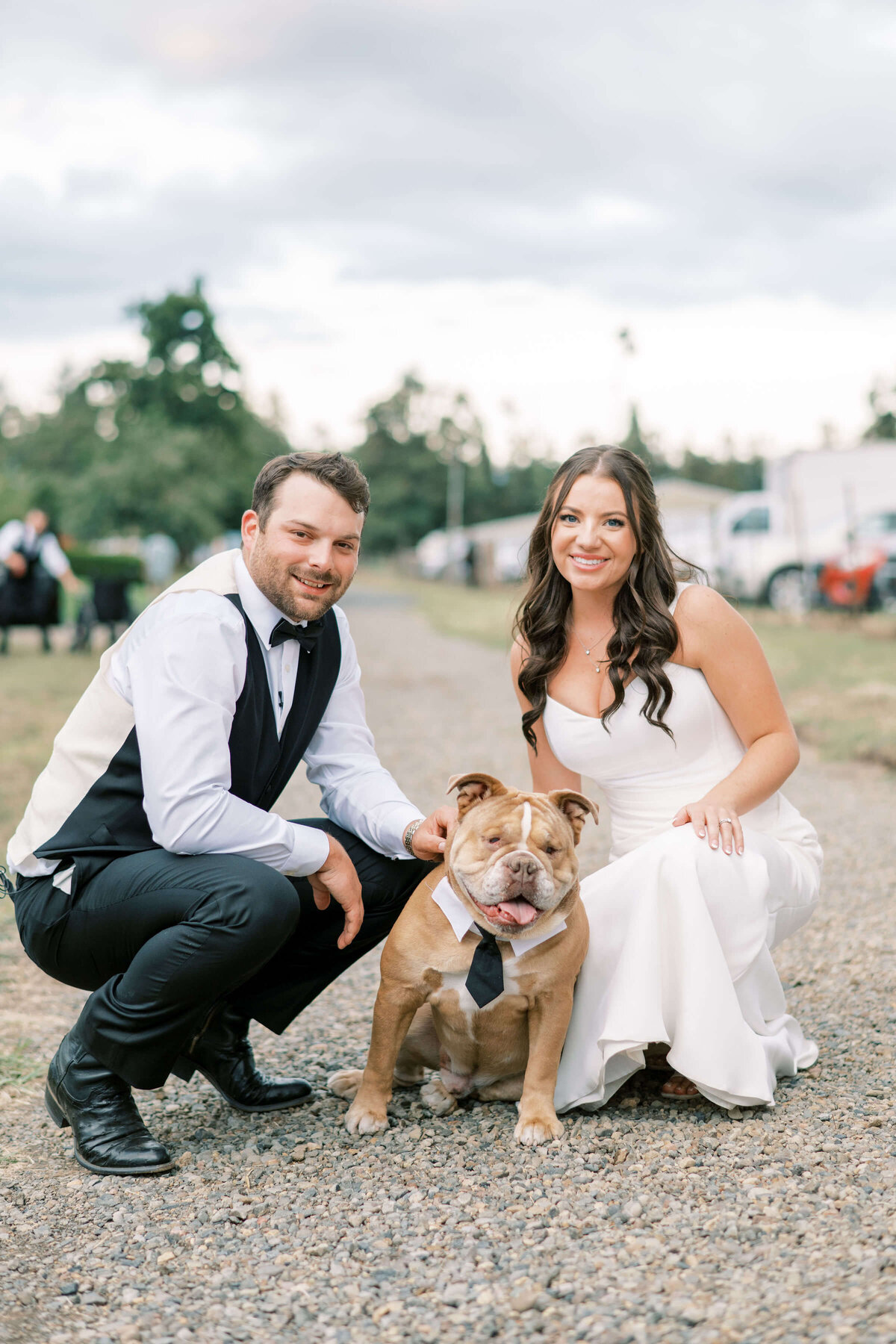 A bride and groom kneel down next to a dog wearing a bow tie and smile and the camera.