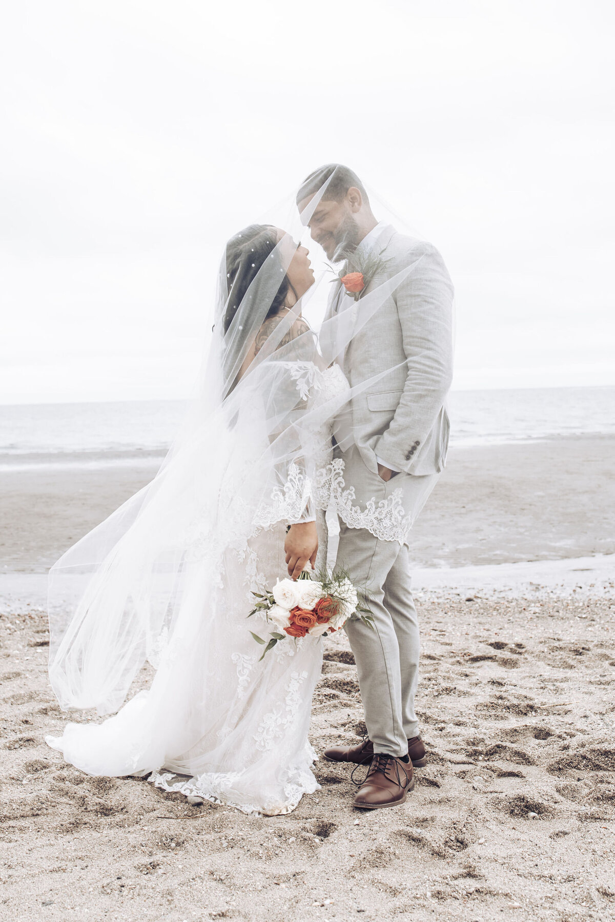 bride and groom standing on the beach wrapped in a veil