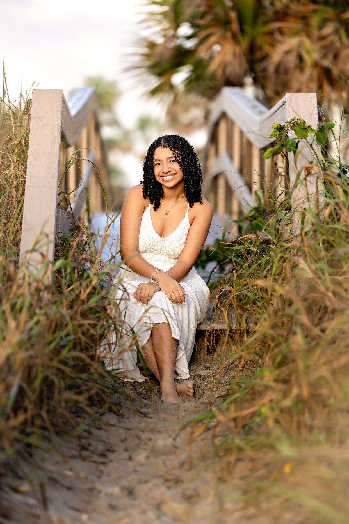 Senior portraits at Cocoa Beach with a teen girl sitting on wooden steps by tall grass, barefoot and glowing