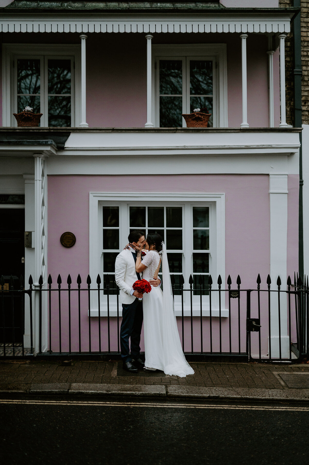 A couple kiss in front of a pink building in London on their wedding day. The groom is wearing a white tux jacket and the bride has a vintage style dress and henna on her hands.