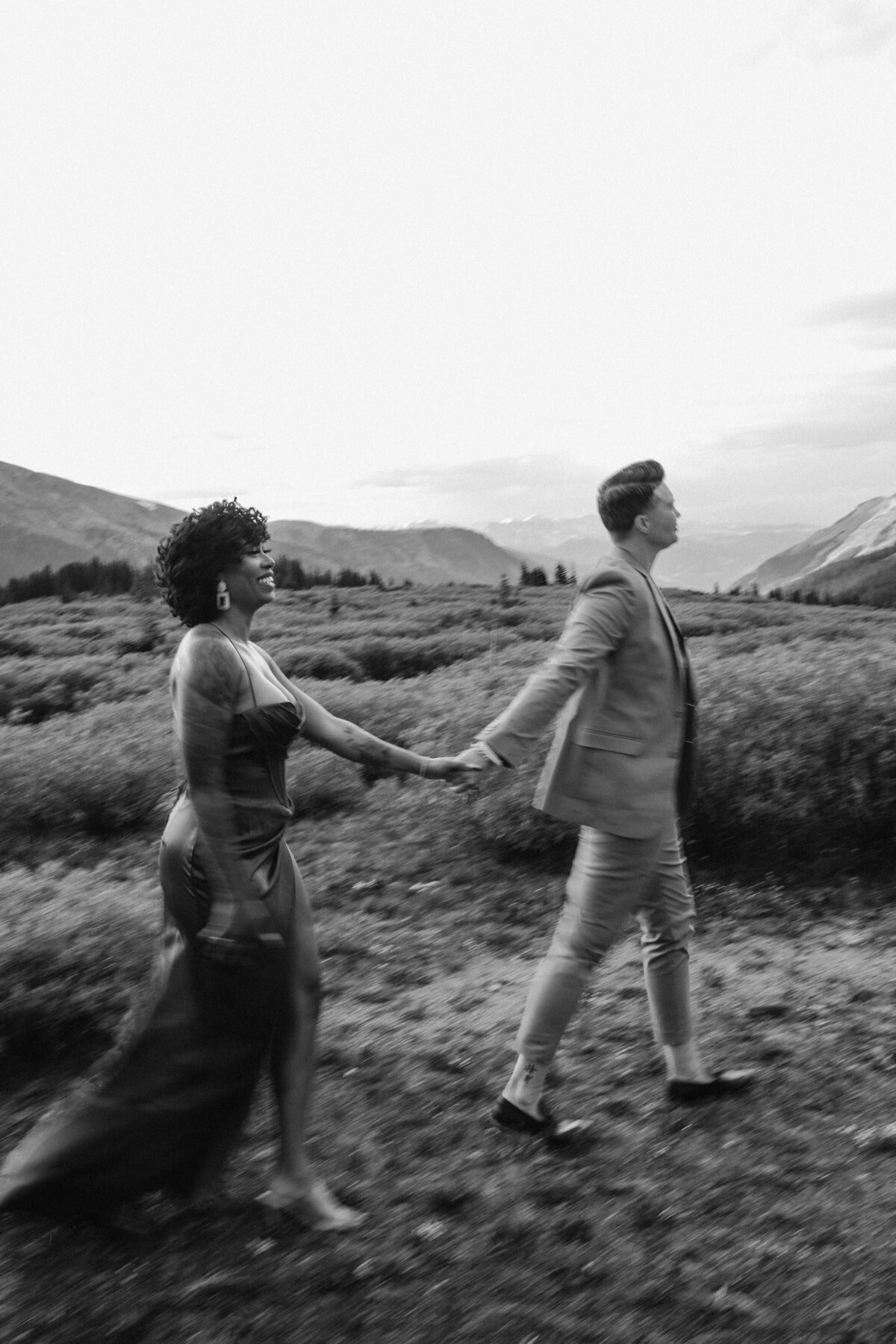 A black and white image capturing a dynamic moment of a couple holding hands and running through a vast field with mountains in the background