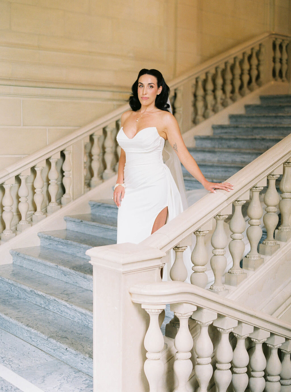 Wedding Photographer RI | A woman in a white gown with a thigh-high slit stands elegantly on a grand staircase with ornate stone railings