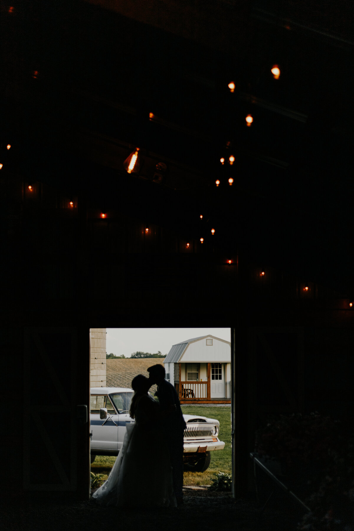 backlit portrait of bride and groom kissing showcasing their vintage car behind them