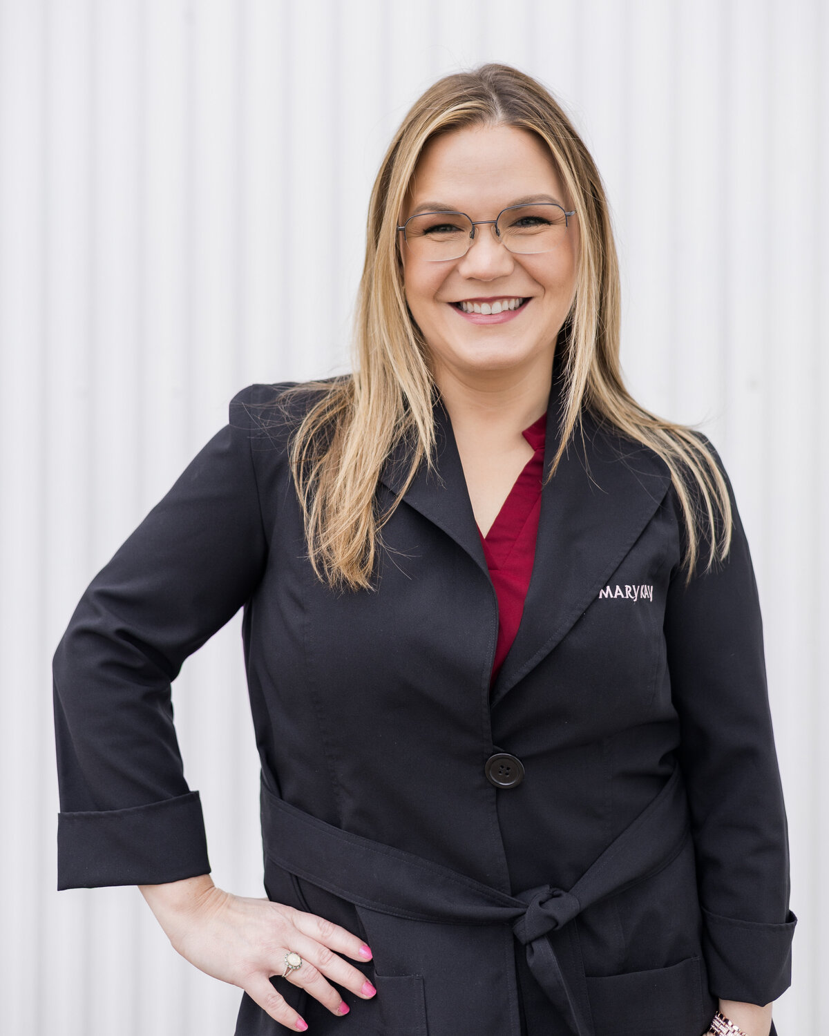 Blonde brunette woman standing in front of a white building, wearing a black Mary Kay jacket with a red shirt under it, wearing glasses, smiling at the camera
