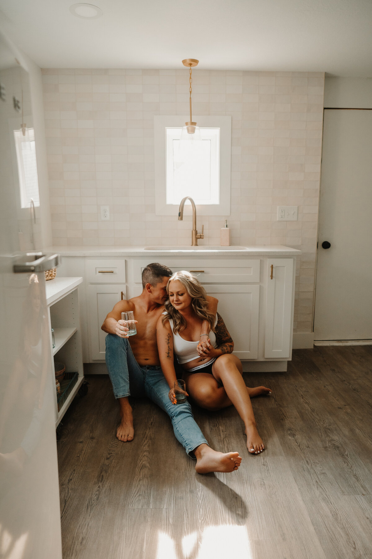 hetero couple sits on white kitchen floor . man with no shirt and jeans on, woman with white crop top and undies on. man has arm around woman and whispers in her ear
