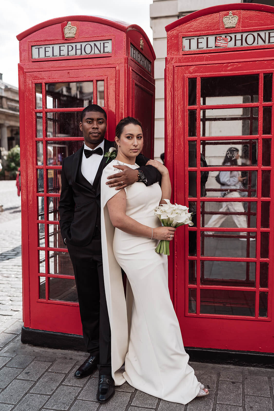 bride wearing a wedding dress with a cape leans against her groom in front of red phone boxes on the streets of london on their wedding day