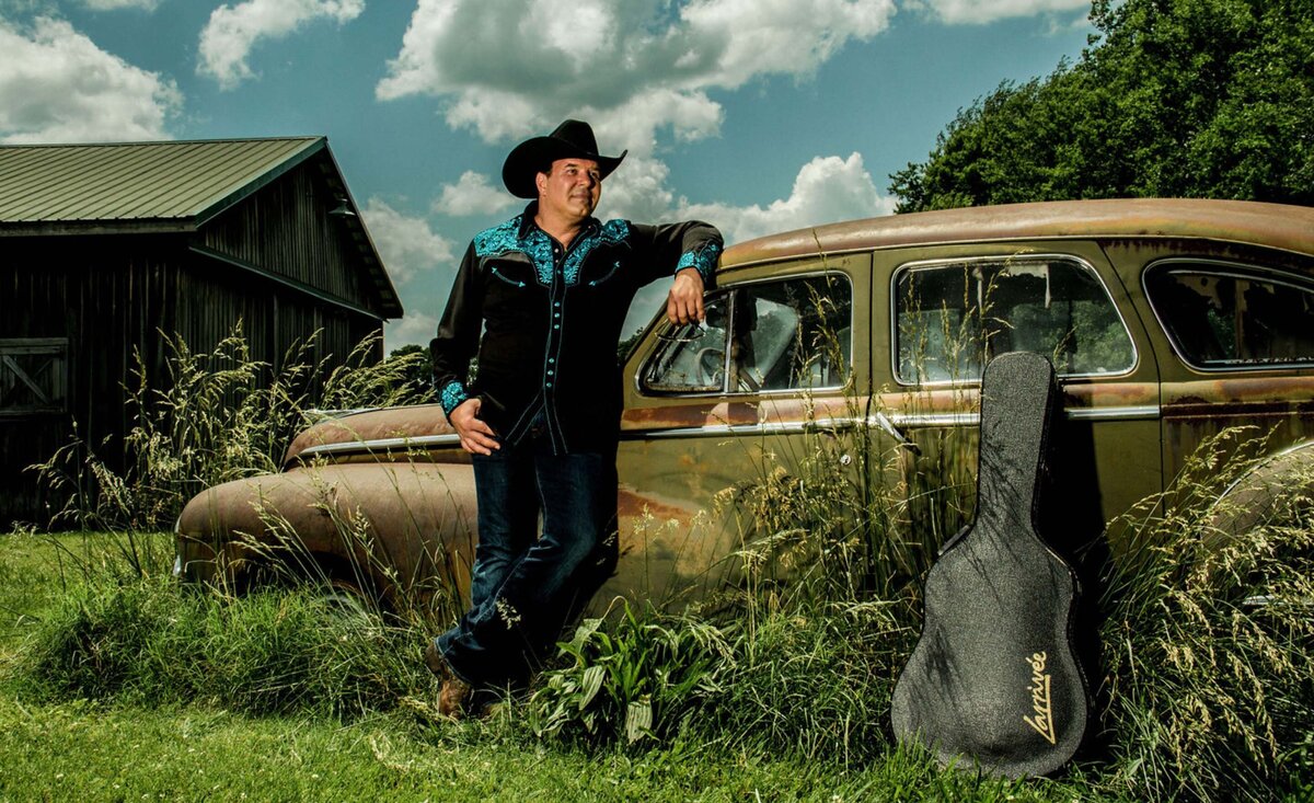 Country musician portrait Ricky Cook wearing black cowboy hat black cowboy shirt leaning against old car with black guitar case under blue sky with puffy clouds