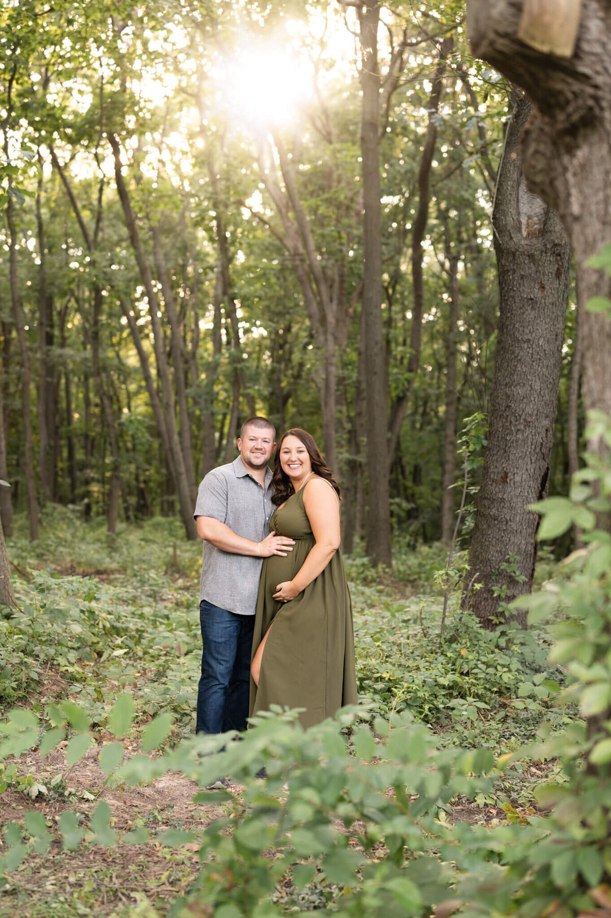 Maternity couple posing under sun positioned behind them in a forest
