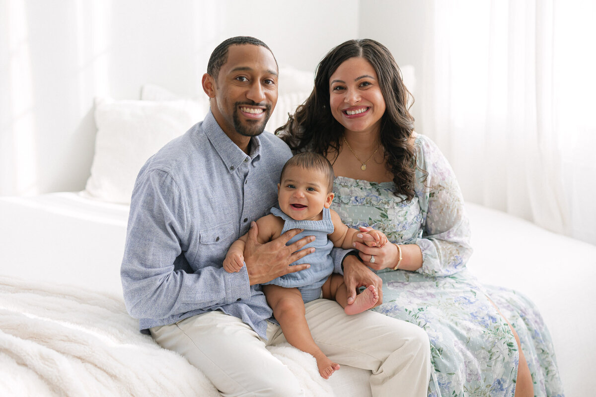 Portrait of a mom, dad and baby sitting on a white bed and smiling at the camera taken by missy marshall photography