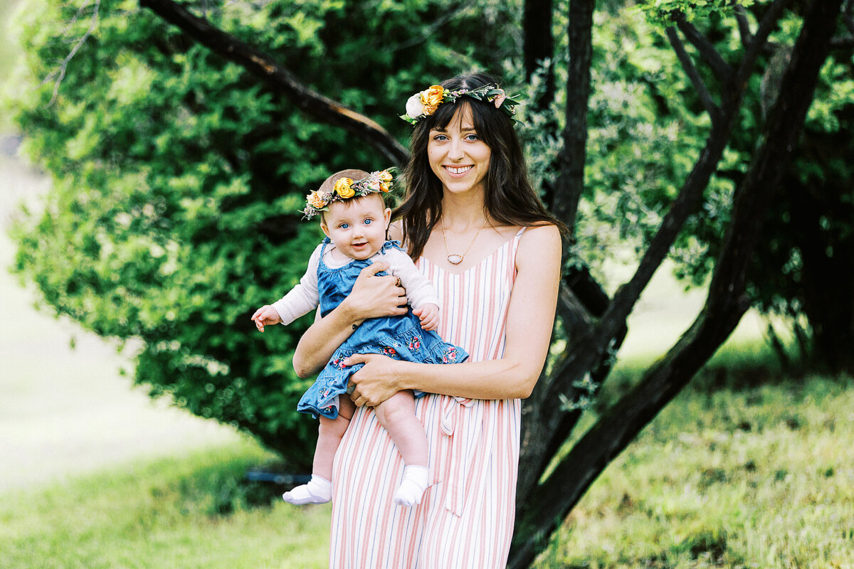a mom in a floral crown holds a healthy baby girl in a floral crown in front of a tree