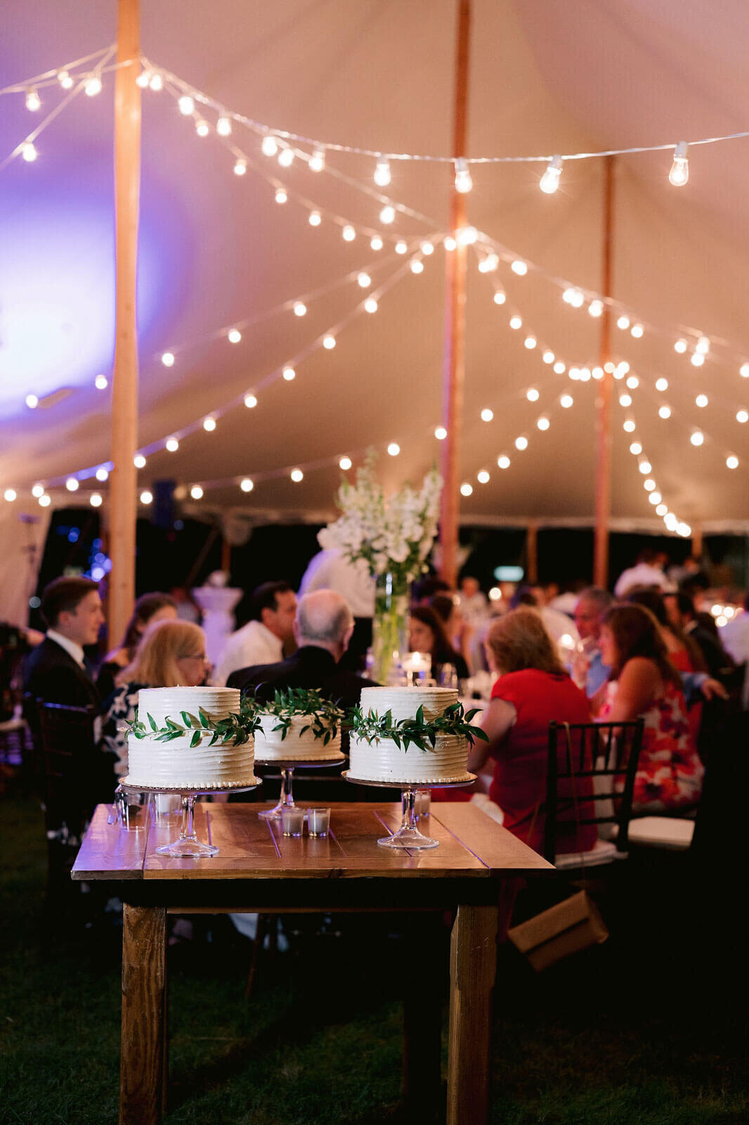 Three white wedding cakes are placed on a wooden table, while the guests are in the background in Cape Cod Summer Tent, MA.