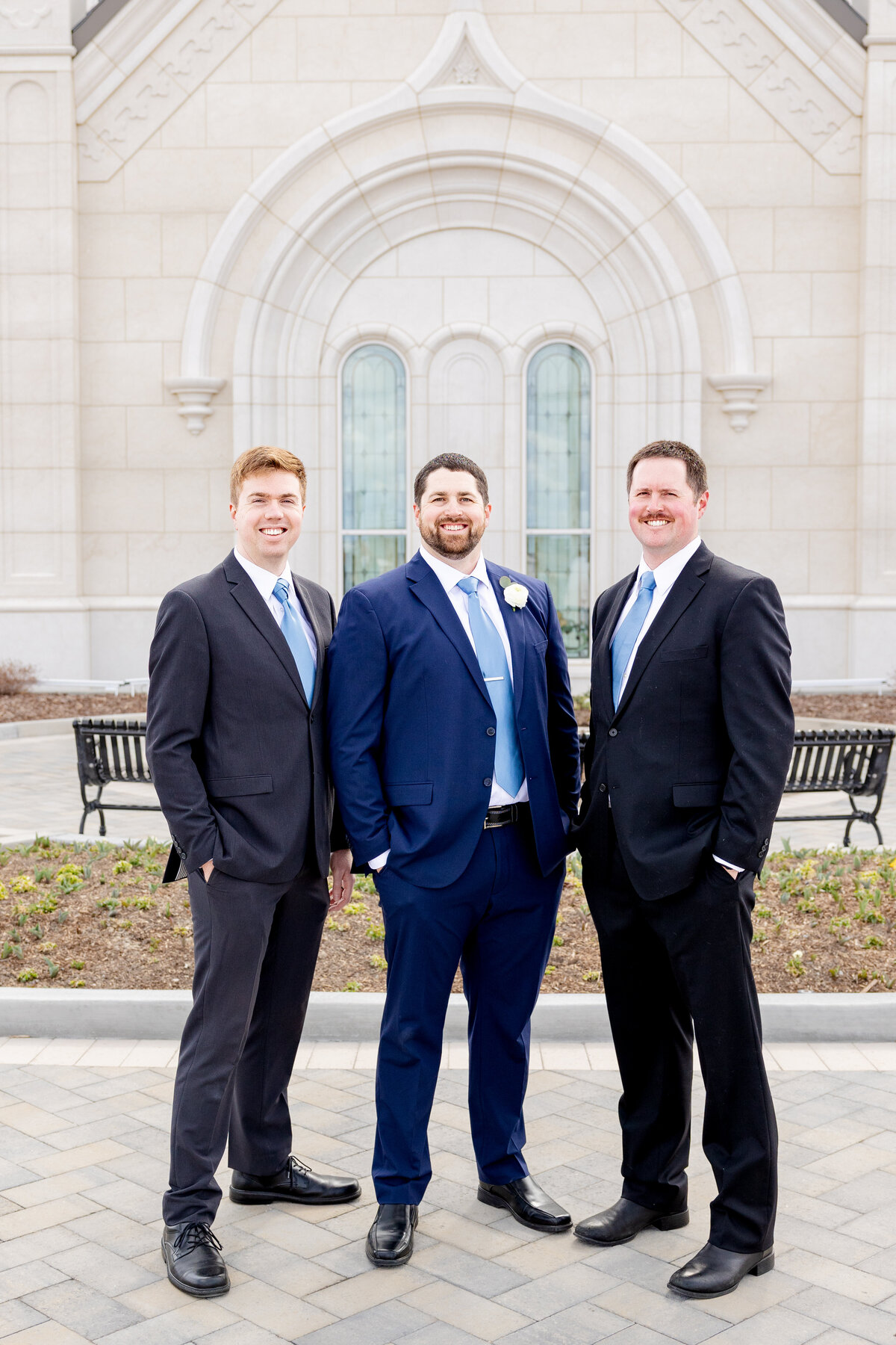 Groom poses proudly with two brothers, all wearing dark suits and blue ties, in front of the decorative windows of the Taylorsville Utah LDS Temple
