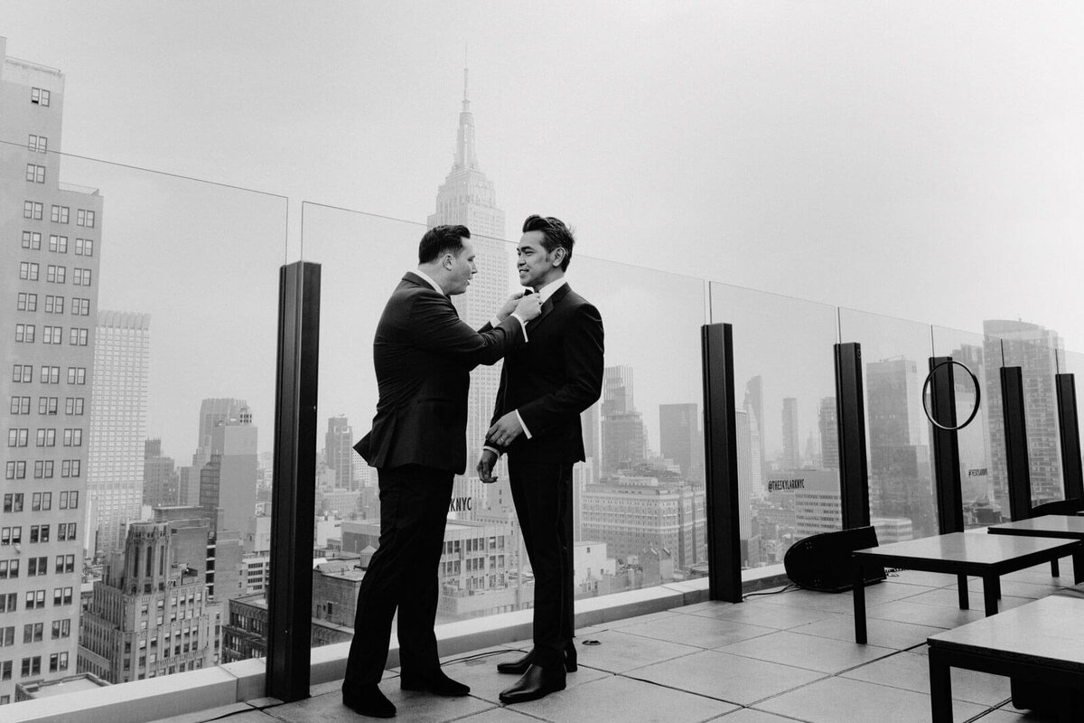 The groom is fixing his groom's bow tie in The Skylark, New York. Buildings in the background. Wedding Image by Jenny Fu Studio