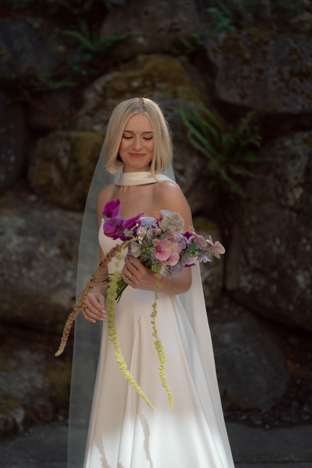 Blonde bride holding colorful wildflower bouquet in sunlight by stone wall