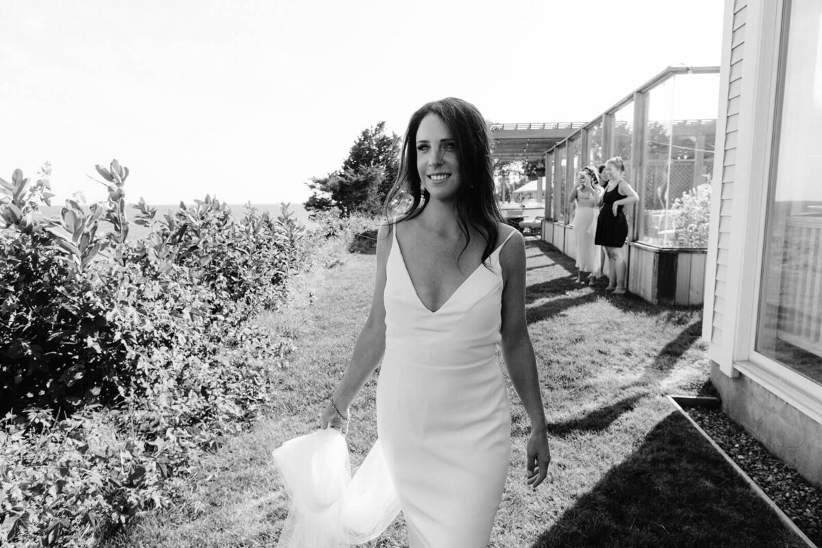 Black and white photo of the bride standing outside Wianno Club, Cape Cod, MA, with plants and trees in the background.