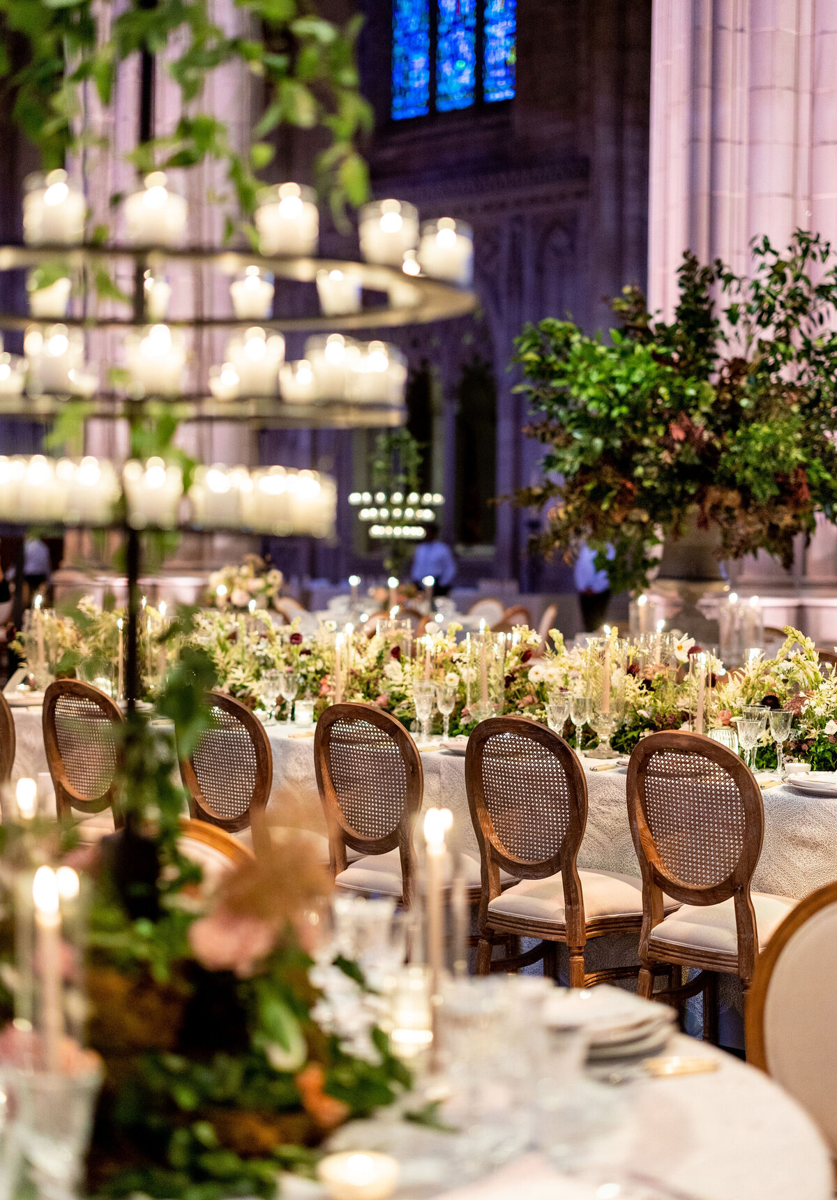 A luxurious dining setup in a grand hall with high ceilings and stained glass windows. Long tables adorned with elegant floral centerpieces, numerous candles, and sophisticated wooden chairs create an opulent ambiance.