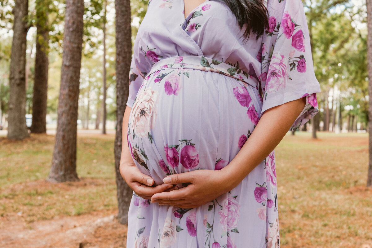 Pregnant woman in a floral dress holding her baby bump during a maternity photoshoot at a serene wooded park. 