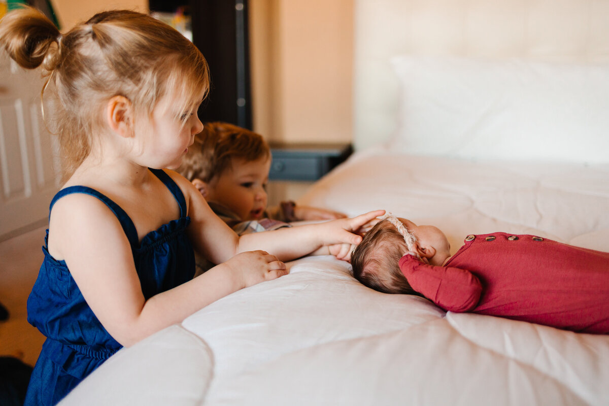 The tender moments with the best maternity photographer in Albuquerque. This heartwarming image features three little siblings attentively watching their newborn sister as she sleeps peacefully on a white bed.