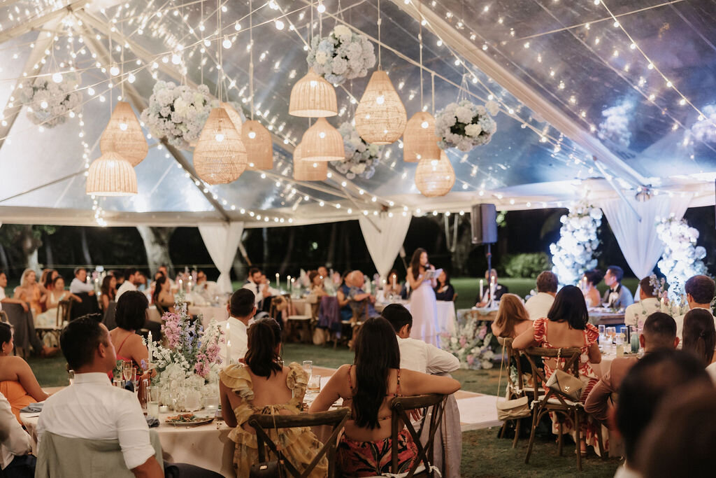 ambient view of lanikuhonua reception with clear top tent, lantern and hydrangea ceiling installation, and wedding guests seated listening to speech
