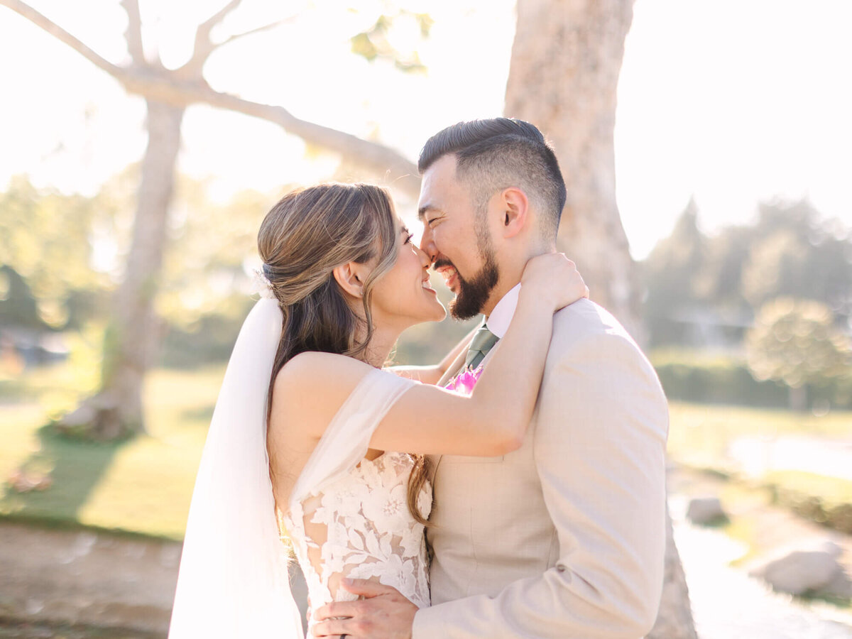 A bride and groom embrace joyfully, touching noses, in a sunlit outdoor setting. The bride wears a lace dress and veil, the groom a light suit.
