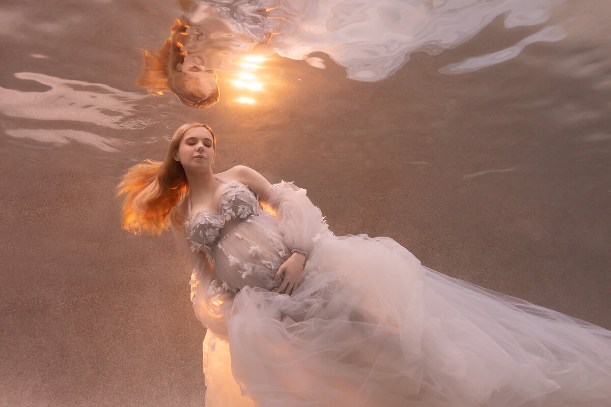 A radiant underwater maternity photo captured by a Dallas underwater maternity photographer. The woman is gracefully floating in a dreamy white gown embellished with floral details. Sunlight glows softly through the water, highlighting her flowing hair and delicate dress, creating an ethereal, golden ambiance.