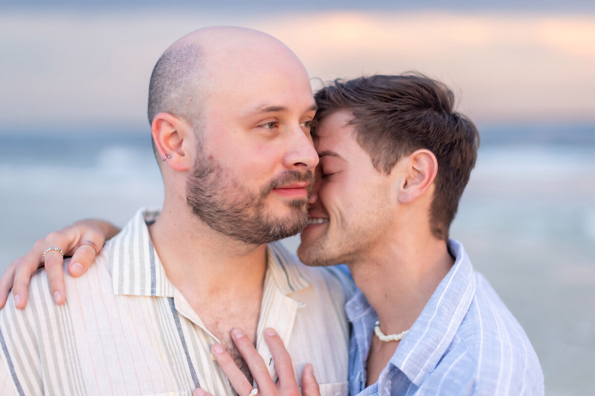 LGBTQ couple hugging close on beach at Deer Lake State park, neither looking at camera while the ring is visible on chest of the proproser.