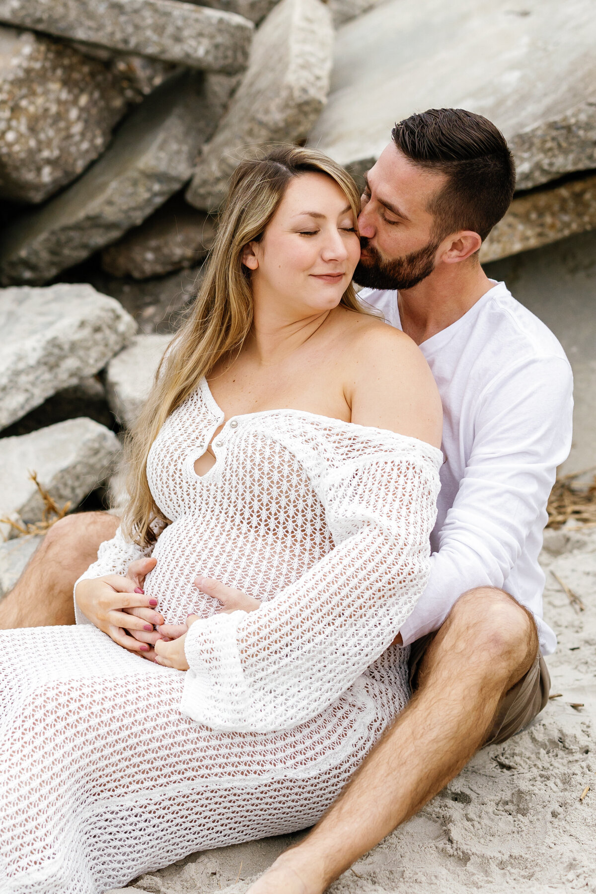 Pregnant couple sitting together on Wilmington beach sand, waves gently lapping nearby