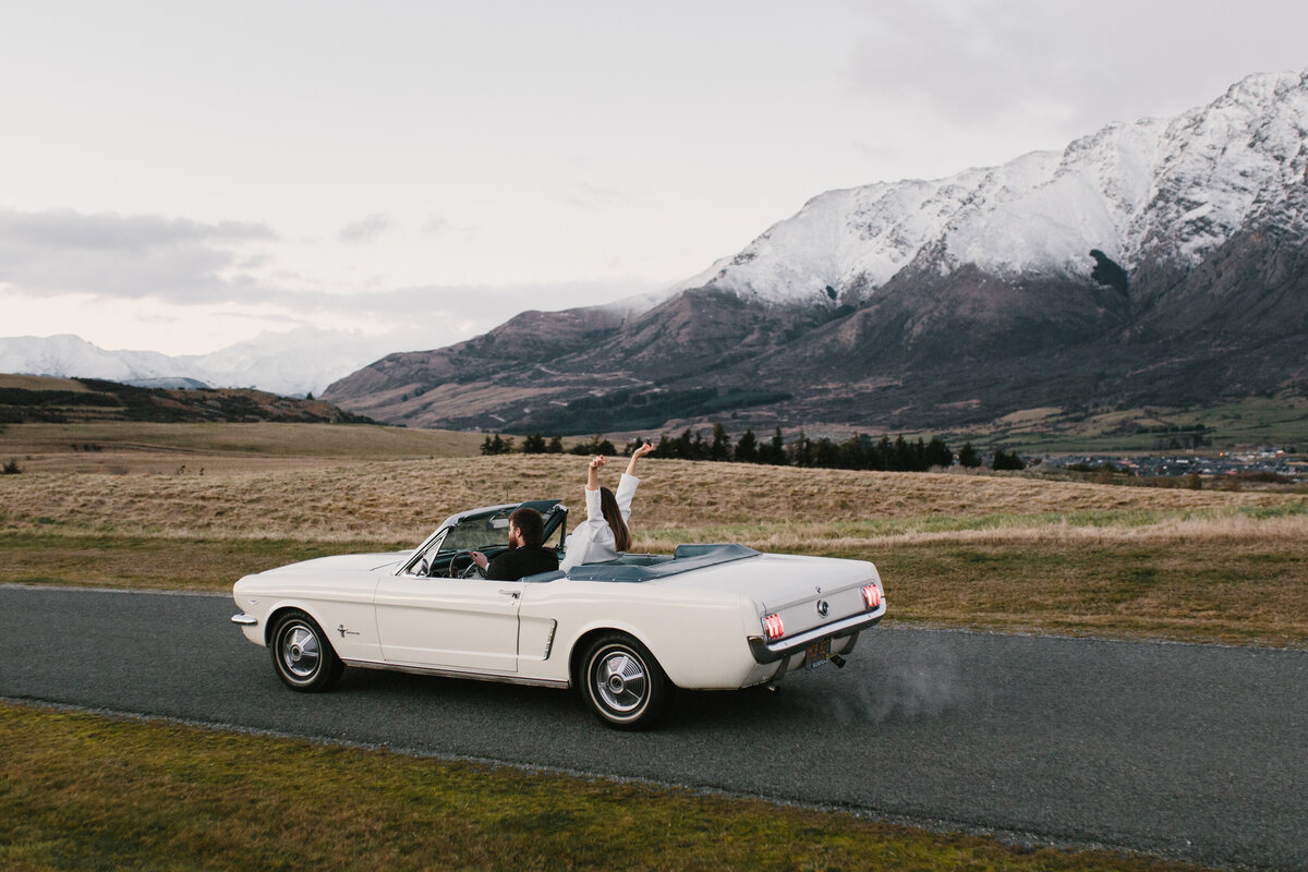 The Lovers Elopement Co - bride and groom drive through Queenstown landscape in New Zealand